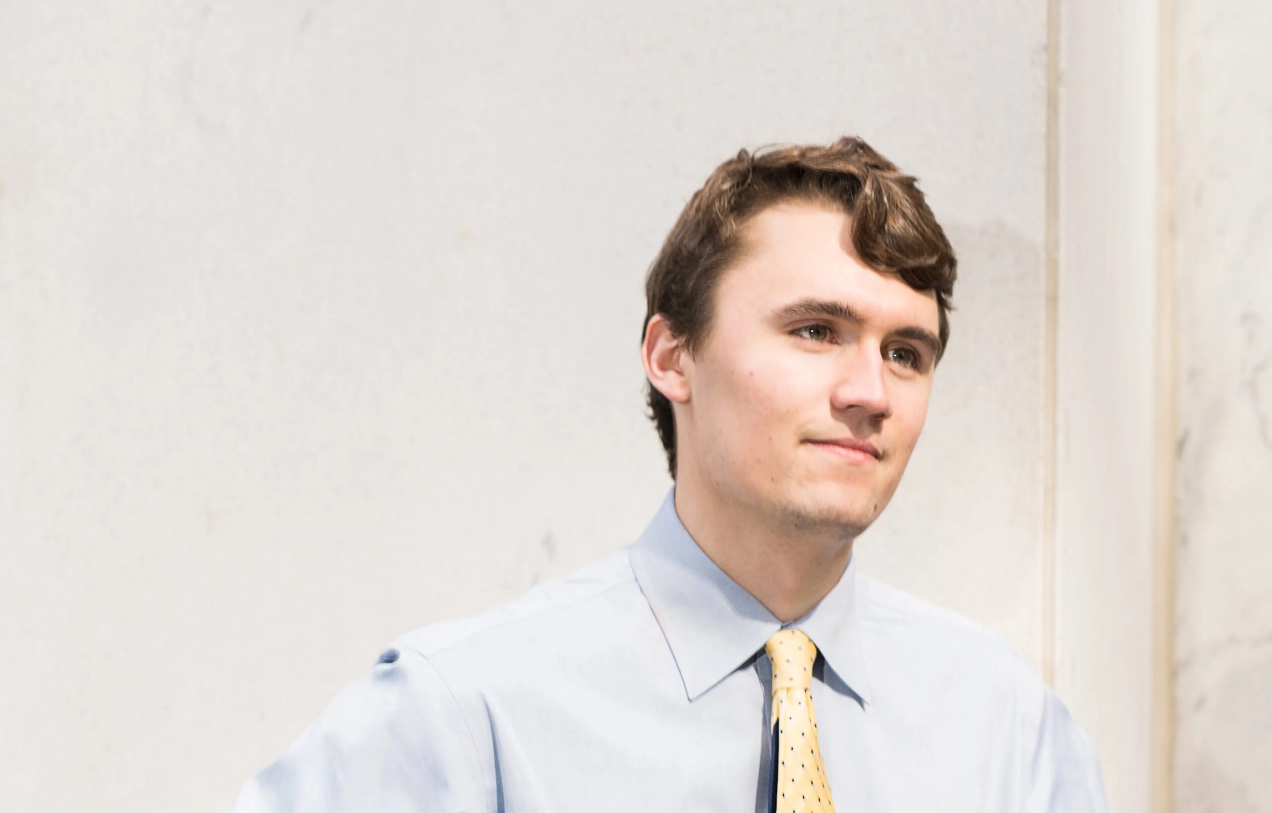 Young Charlie Kirk wearing a light blue shirt and yellow tie, standing against a light-colored wall with a composed expression.