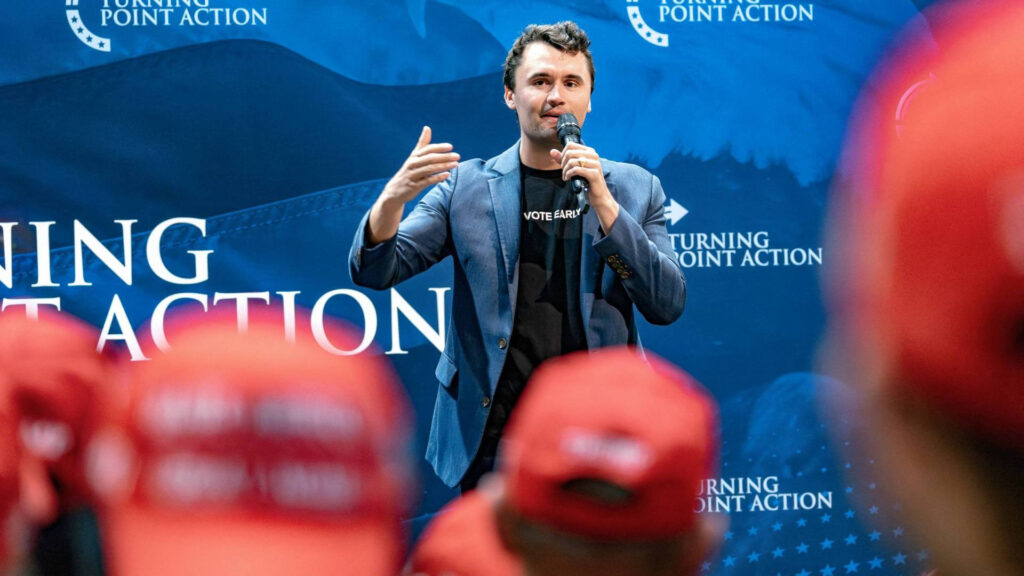 Charlie Kirk speaking into a microphone at a Turning Point Action event, standing in front of a blue backdrop with red-hatted attendees in the foreground.