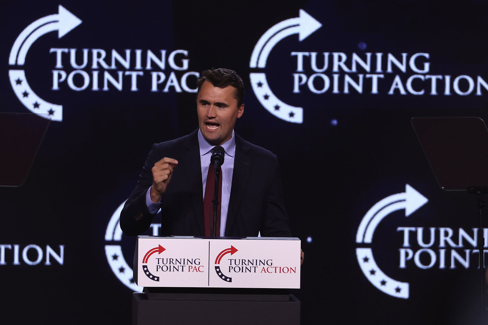 Charlie Kirk passionately speaking at a podium with Turning Point Action and Turning Point PAC logos in the background.
