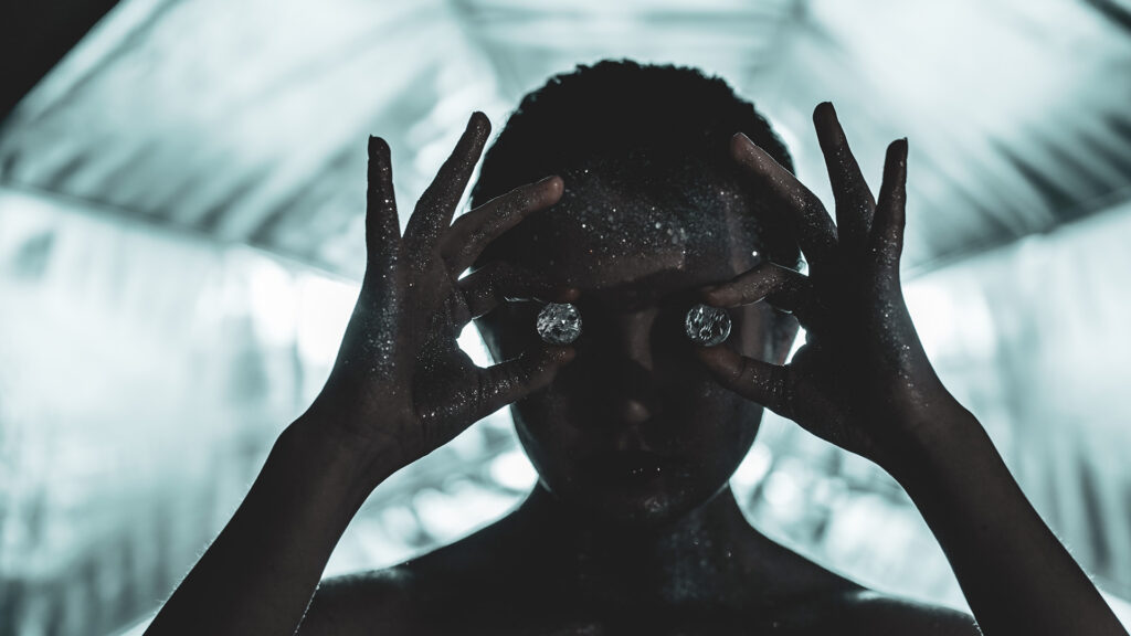 Silhouette of a person covered in glitter holding two round diamonds over their eyes, standing in a cool, backlit tunnel.