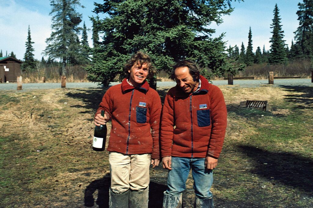 Two men in red Patagonia fleece jackets standing outdoors with muddy pants, one holding a champagne bottle, both laughing under a tree.