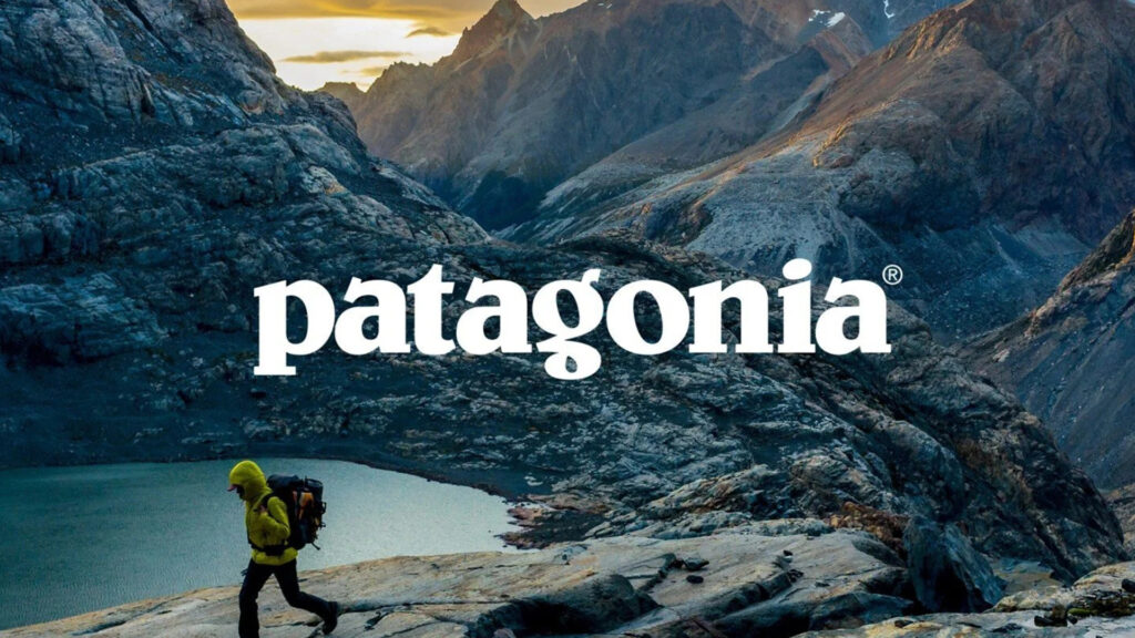 A hiker in a yellow jacket treks through rocky mountain terrain near a lake with the Patagonia logo overlaid in white.