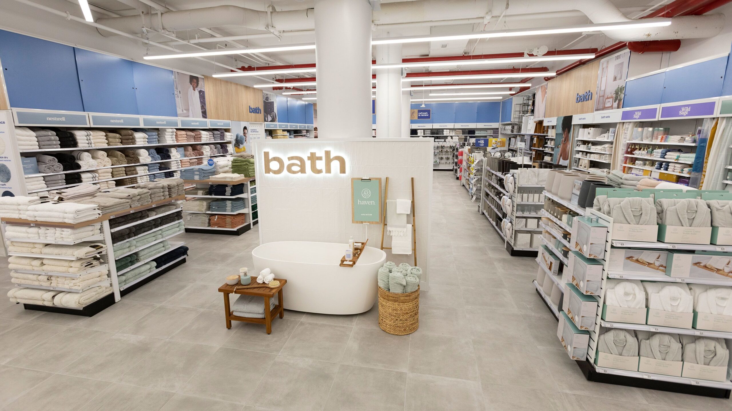 Well-lit and organized bath section of a Bed Bath & Beyond store with neatly stacked towels, robes, and a freestanding tub display.