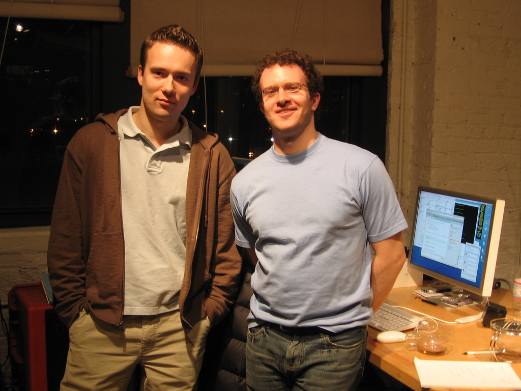 Basecamp founders Jason Fried and David Heinemeier Hansson standing confidently in front of a yellow background with the Basecamp logo.