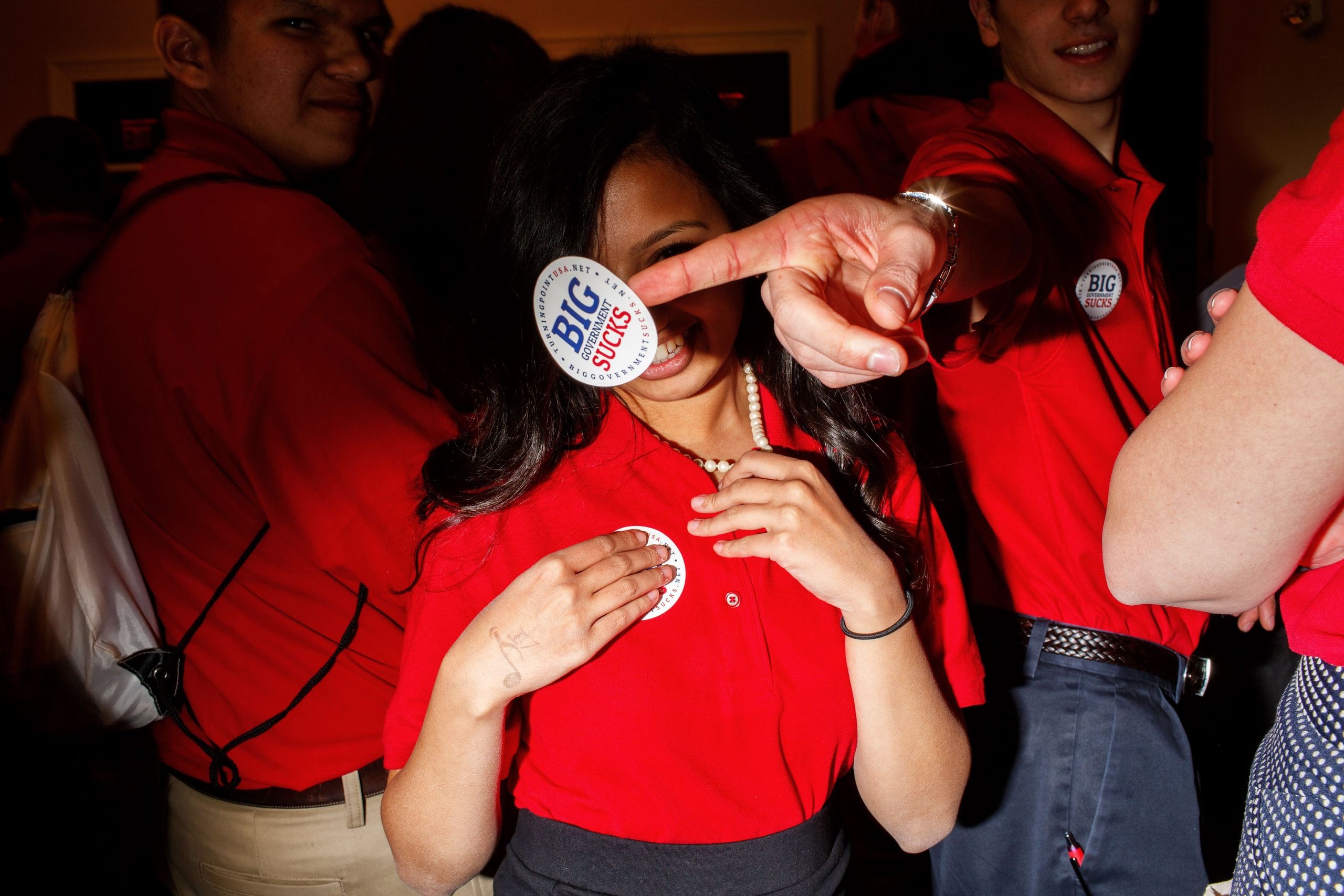 Young people in red shirts at a Turning Point USA event, with one person holding a "Big Government Sucks" sticker in front of the camera.