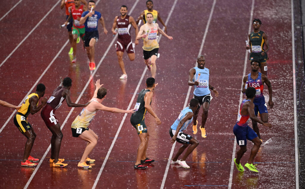 Letsile Tebogo  hands the baton to Bayapo Ndori  during the 4x400m relay at the World Athletics Championships Tokyo 2025, with multiple teams exchanging in the rain.