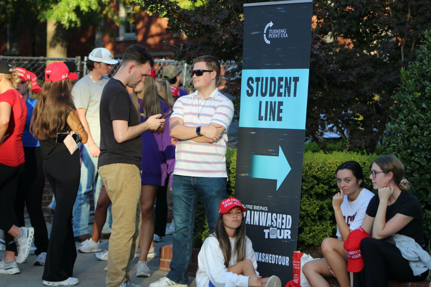 Students gathered near a “Student Line” sign at a Turning Point USA event, with some wearing red MAGA hats and others sitting near the banner reading “Culture Being Brainwashed Tour.”