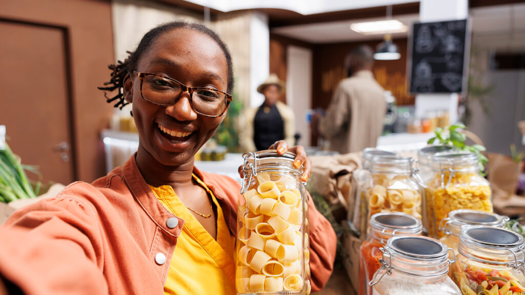 Sustainable Products Modern-shop – Tumisang Bogwasi Smiling person in glasses holds a jar of large pasta shapes while taking a selfie in a bright refill shop with rows of glass jars and plants in the background.