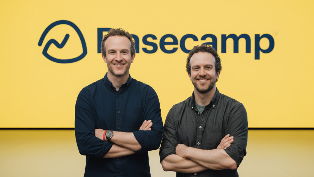 Jason Fried and David Heinemeier Hansson standing in front of a bright yellow wall with the Basecamp logo, arms folded and smiling.
