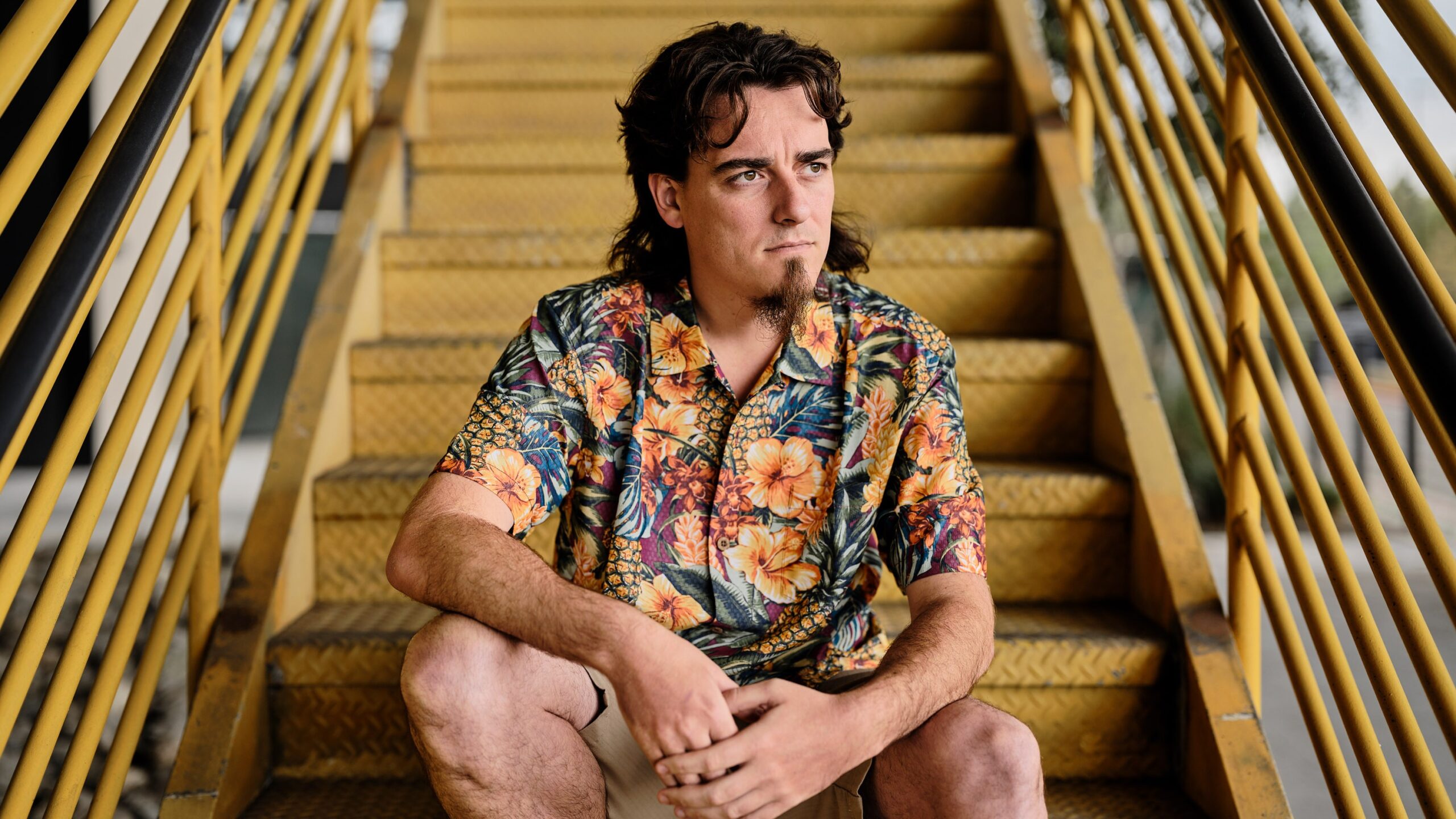 Palmer Luckey, founder of Anduril, seated on yellow metal stairs wearing a colorful floral shirt, looking off thoughtfully.