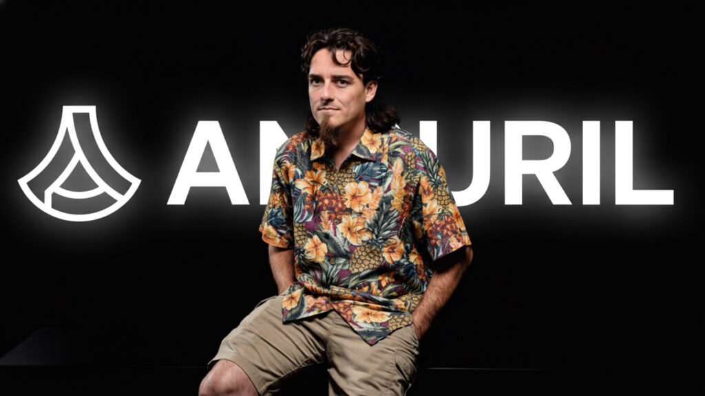 Palmer Luckey wearing a colorful floral shirt and cargo shorts, seated in front of a glowing Anduril logo on a black background.