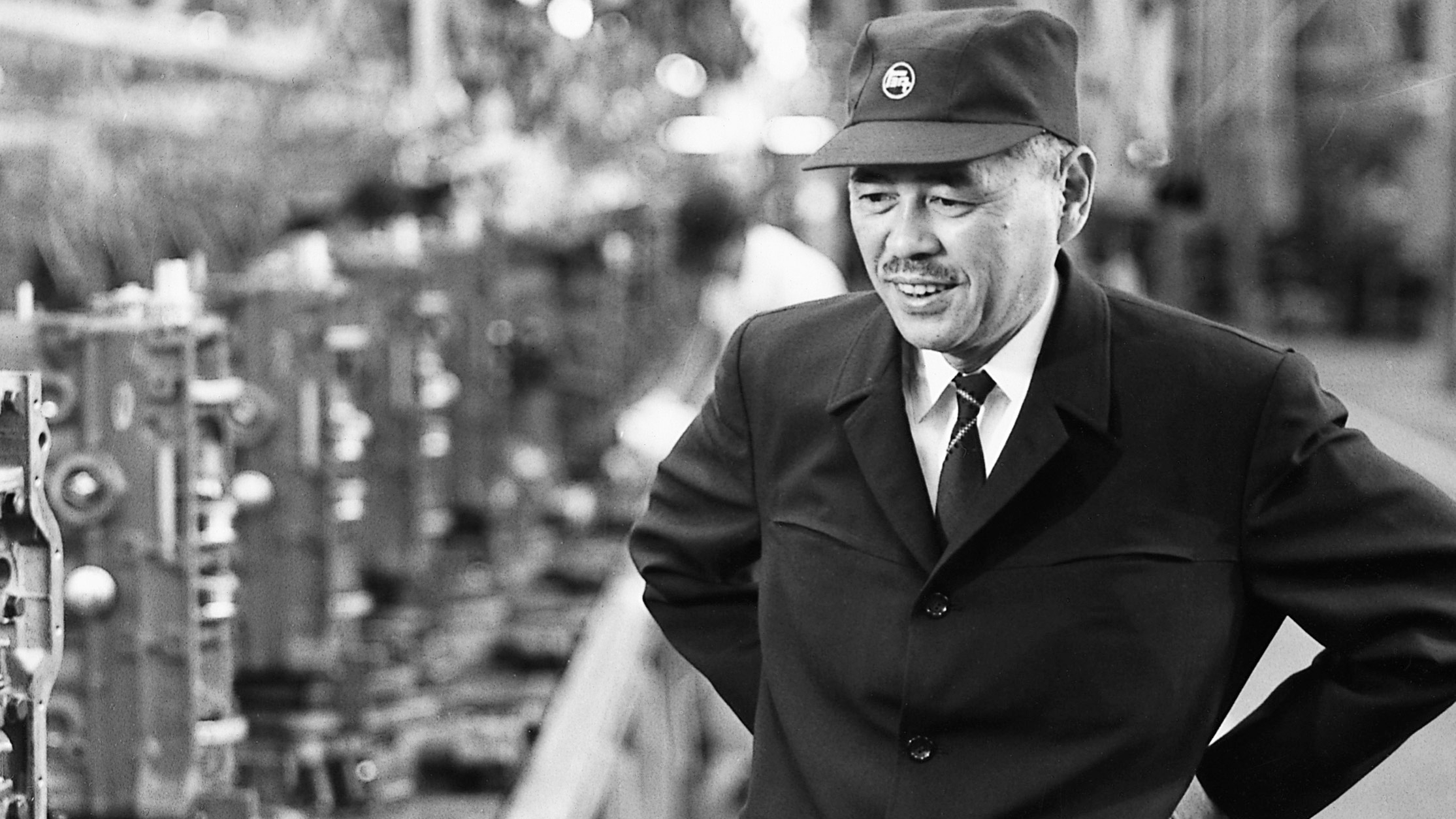 Taiichi Ohno Father of the Toyota Production System – Tumisang Bogwasi Taiichi Ohno smiling and standing on a factory floor in uniform and cap, surrounded by industrial machinery
