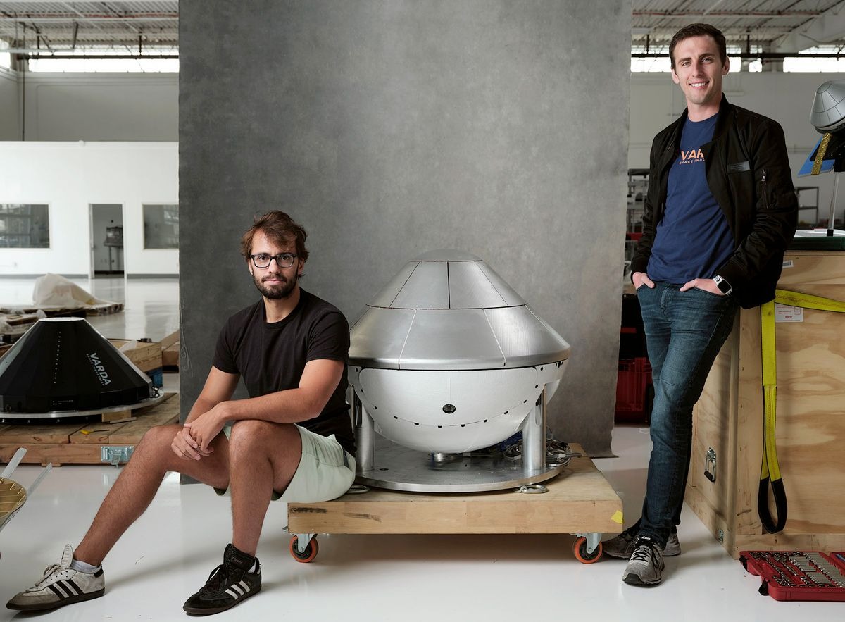Delian Asparouhov and Will Bruey, co‑founders of Varda, standing and seated beside a spacecraft manufacturing prototype inside an industrial workspace.
