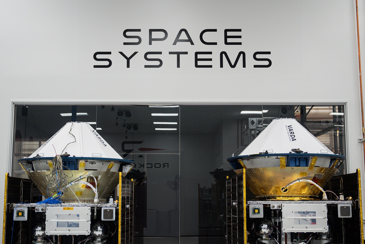 Two Varda reentry capsules positioned inside a Space Systems lab, showing heat shield assemblies, avionics hardware, and structural components under controlled testing conditions.